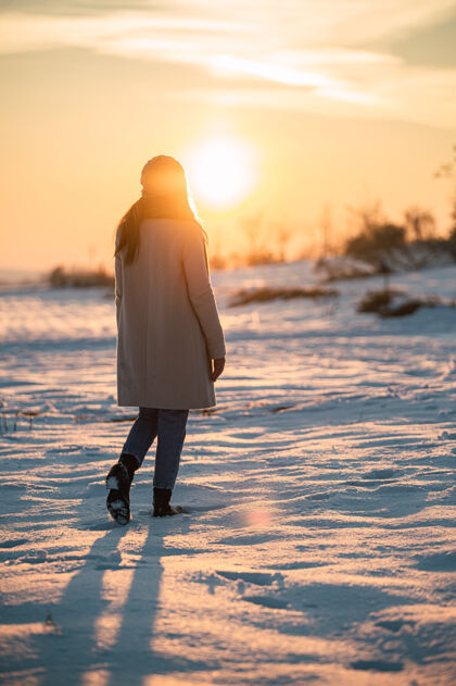 匿名一个穿着暖和衣服的女人独自站在雪白的草地上 欣赏冬日乡间如画的日落阳光背景风景