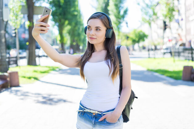 耳机Instagram影响者小工具设备学生牛仔服装概念美丽迷人美丽美丽自信的女士的肖像在公园镇拍摄自拍周末旅游游客手机走路手机
