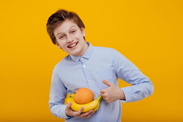 秀手里拿着水果 香蕉和橘子 穿着蓝色衬衫 孤零零地站在黄色的墙上的快乐少年新鲜香蕉英俊