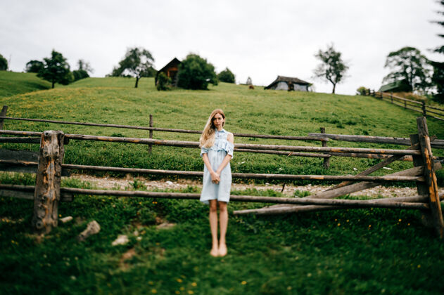 木头年轻漂亮的金发碧眼的女孩 穿着蓝色的裙子 在乡村的篱笆边摆姿势女人优雅姿势