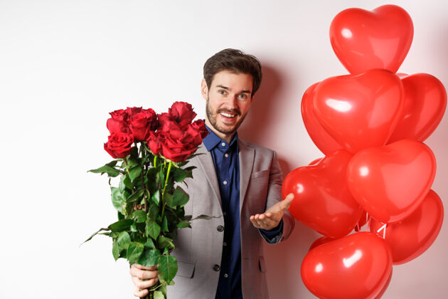 伴侣穿西装的浪漫男人说情人节快乐 送红玫瑰给你 还指着你的手 站在白色背景上 心形气球礼物情人节帅哥玫瑰