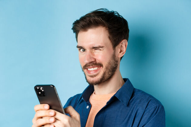 发型有胡子的英俊年轻人的肖像 对着相机眨眼微笑 用智能手机 站在蓝色背景上朋友站立科技