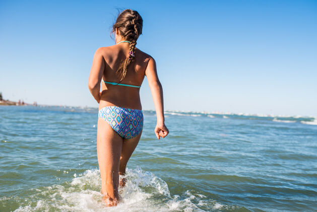 天空正面小女孩在喧闹的海浪中在水面上奔跑的后视图欢笑玩耍年轻