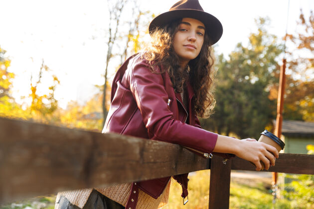 欢乐美丽的年轻女子戴着帽子 斜靠在秋日公园的木桥上 端着一杯外卖咖啡温暖概念树叶