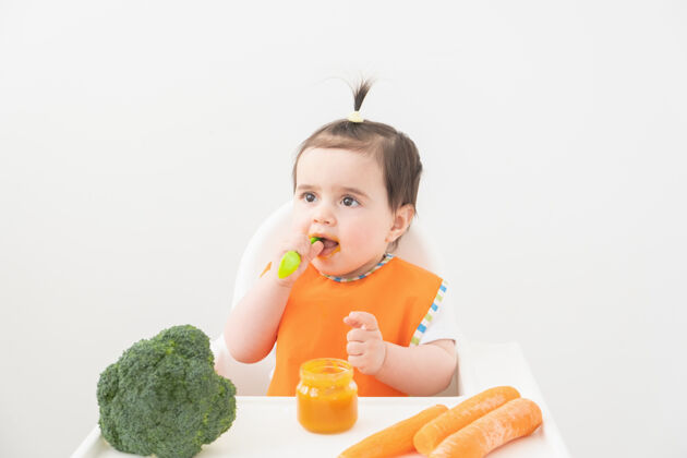 花椰菜穿着橙色围嘴的女婴坐在儿童椅上吃白色背景的蔬菜泥餐饮食护理
