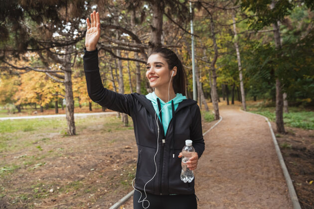 积极微笑的健身女 戴着耳机听音乐 站在公园里拿着一瓶水饮用水白种人休息