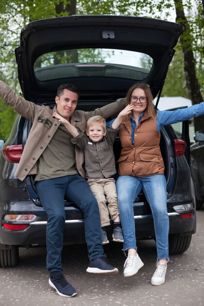 父母幸福的夫妇和他们的小儿子在开放的越野车后备箱在停车场停车道路孩子