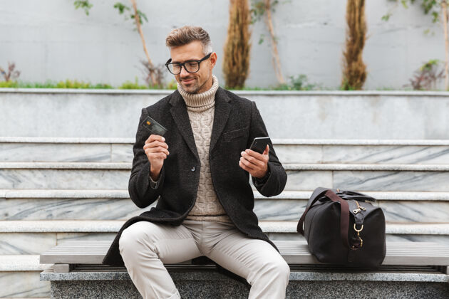 支付帅哥穿着夹克坐在户外用手机和信用卡上网购物支付客户应用程序