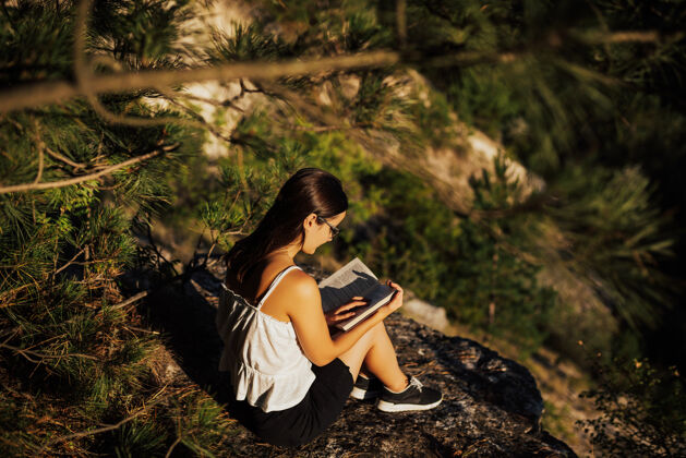 自由年轻的姑娘戴着眼镜坐在山上的岩石上看书 在宁静阳光明媚的夏日里 充满了温暖的阳光年轻举行华丽