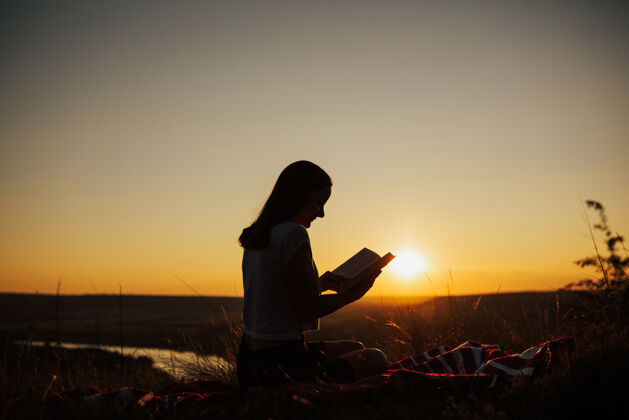 旅行日落时分 一个年轻漂亮的女人坐在格子布上 仔细地盯着打开的书自由环境阅读