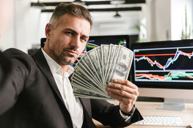 吸引力富商30多岁穿着西装拿着钱扇在办公室工作的图像 电脑上有图形和图表工作统计图形