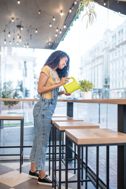 干净关心 植物微笑穿着休闲服和运动鞋的年轻女子在咖啡馆浇灌植物忙碌工作日咖啡馆