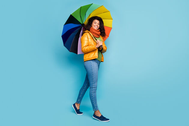 女深色皮肤的女士带着蓝色背景的彩虹色雨伞行走蓝色背景外套牛仔裤