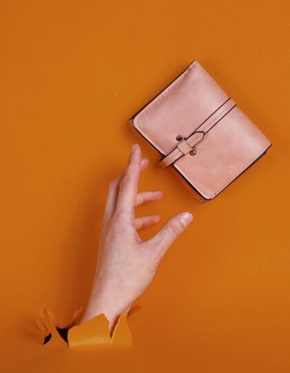 创意女性手拿粉色钱包穿过撕破的橙色纸简约创意时尚理念年轻内想法