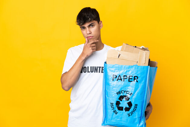 志愿者男人拿着一个装满废纸的回收袋 在孤立的黄色背景下思考着思考解决方案困惑