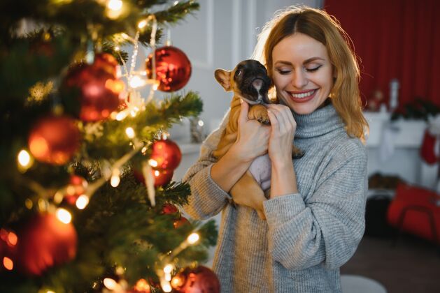 年轻美丽的年轻女子带着一只小狗在圣诞庆典的背景下拥抱欢呼室内
