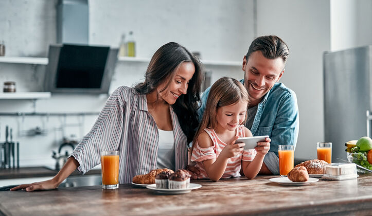 早餐中国的幸福家庭观厨房可爱小女孩和她美丽的父母正在用智能手机 微笑着准备美味的早餐妻子自拍女儿