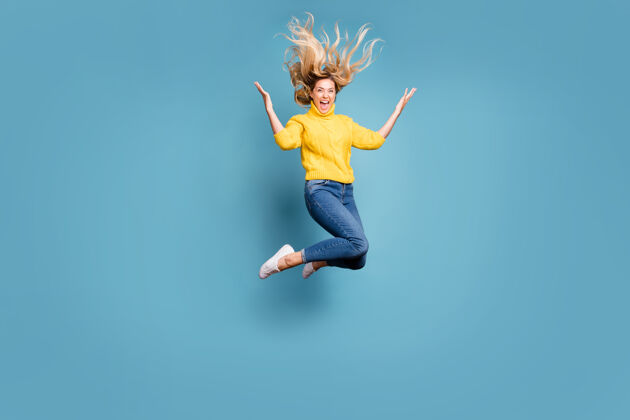 跳投疯狂女士跳高享受秋风发型飞扬开朗人穿针织黄色套头衫牛仔裤隔离蓝色墙壁的全身照片女人空气胜利