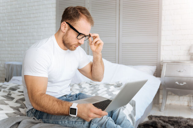程序员时间到了工作快乐专注的大胡子男人坐在家里的床上 一边用笔记本电脑一边表达专注和工作科技当代电脑