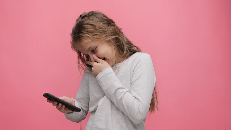 童年快乐可爱的小女孩手持智能手机 站在粉色背景上大笑的照片休闲兴趣活动