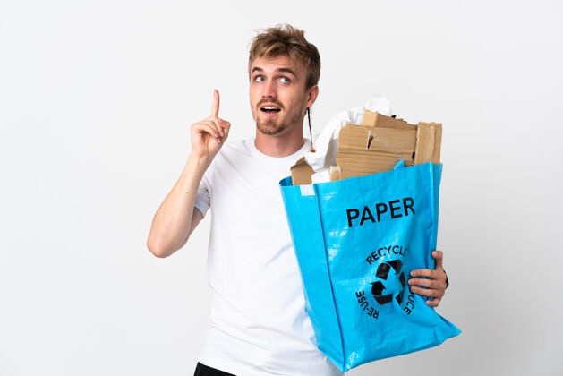 生态年轻的金发男子拿着一个装满纸的回收袋 在白色的纸上进行回收 意图实现解决方案的同时举起一根手指环境环境志愿者