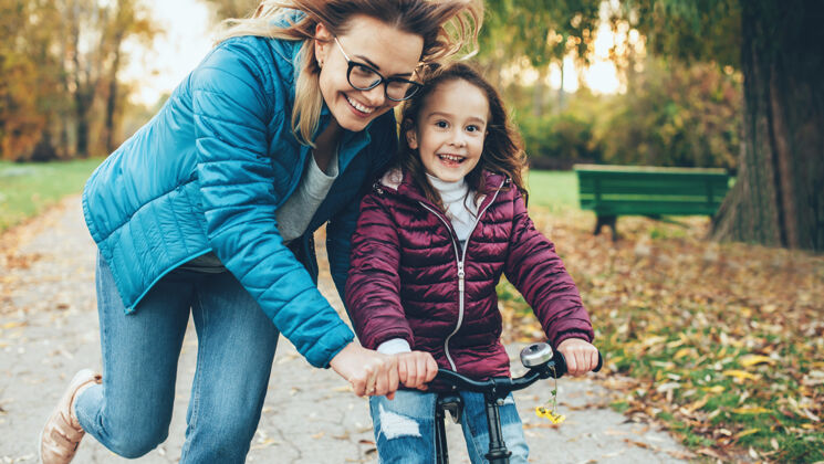 情绪戴着眼镜的可爱妈妈试图教女儿在公园里骑自行车笑城市户外