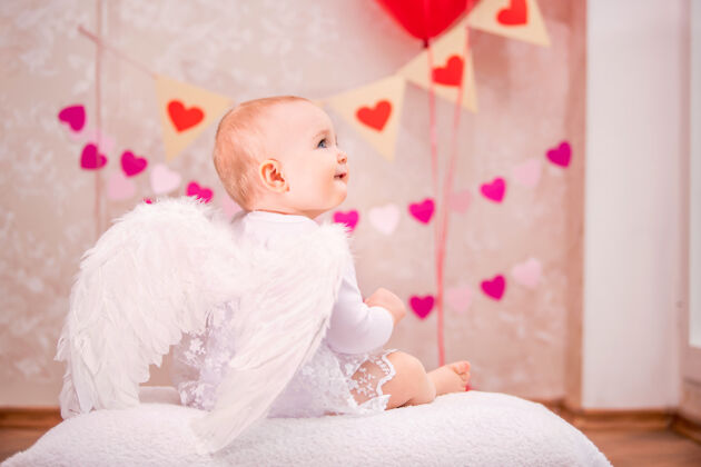 孩子带着白色羽毛翅膀的女婴坐在白色柔软的枕头上 从后面看是情人节的象征人粉色宝贝