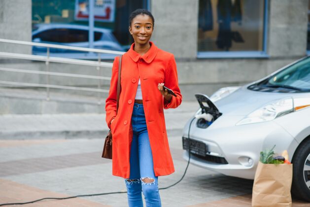搜索年轻女子站在电动车旁边 看着摄像机共享持有插座