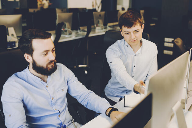 发展一个穿白衬衫的年轻人一边指着电脑显示器 一边帮留着胡子的同事做项目办公室男同事们一起在办公室工作计算机协作设备