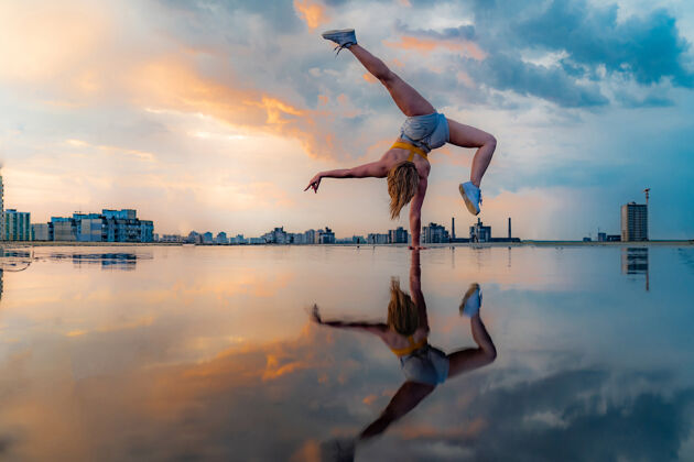 自由女子体操运动员站在一只手上 并保持平衡 在戏剧性的日落与反映在水惊人的云彩城市瑜伽天空