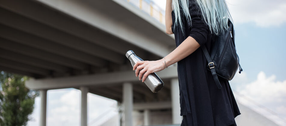 女士年轻的金发女孩的画像 穿着黑色的衣服 背着背包 拿着钢瓶 在现代桥下闪亮温度清洁