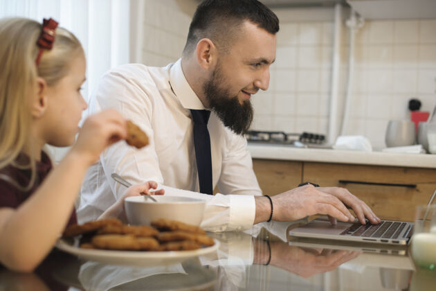 糕点一位留着胡须的年轻父亲的侧视图 他穿着西装去上班 看着厨房里的笔记本电脑 而他的女儿正在吃饼干男人巧克力笔记本电脑