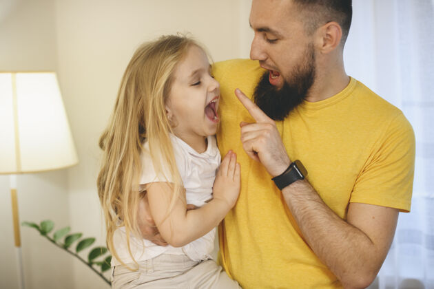 手表小胡子的父亲和他的小女儿在家里玩耍早上好可爱金发碧眼的小女孩一边玩乐一边咬爸爸的手指表情爸爸小