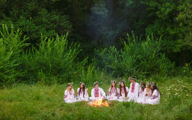 阳光仲夏年轻穿着斯拉夫服装的人们坐在火堆附近的树林里时尚神圣仪式
