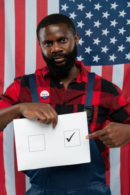 非洲人后裔当代年轻的非洲裔美国修理工指着选票上的一个正方形的滴答声 同时站在美国国旗的对面国旗爱国选举