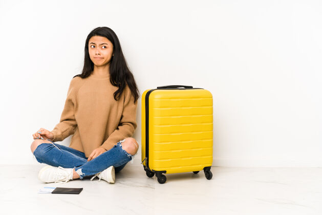 嘴唇坐在地板上拿着手提箱的年轻中国女旅行家感到困惑 怀疑和不确定怀疑皱褶旅行