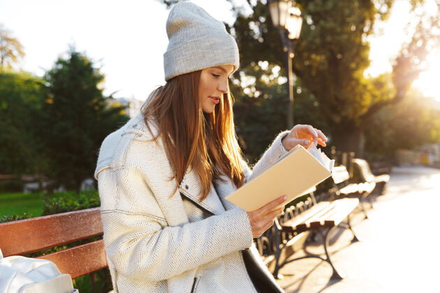 时尚一个穿着秋衣戴秋帽的漂亮年轻女子坐在户外的长凳上看书阅读吸引温暖