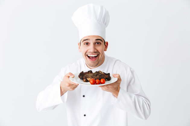 食谱开朗的厨师穿着统一的胡椒粉在一个盘子上煮牛排 隔着白墙热烧烤男性