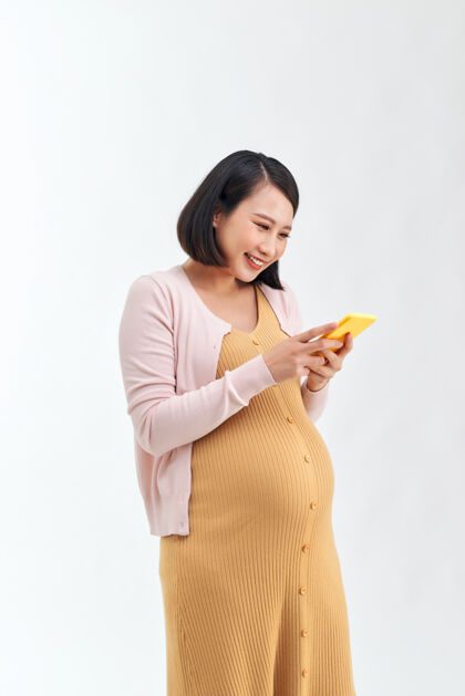 成人一个快乐的年轻孕妇在白墙背景下用手机摆姿势中国怀孕站