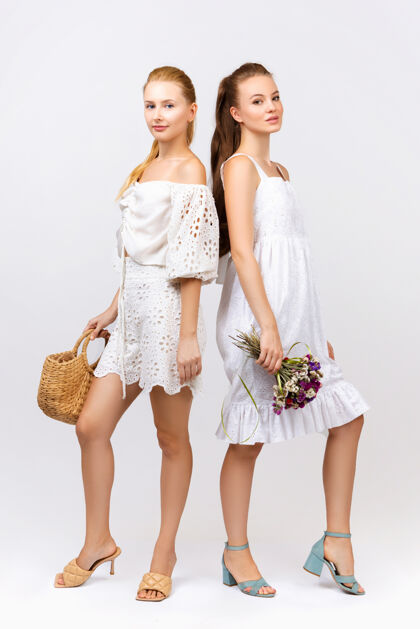 女人两个穿着白色连衣裙 手拿手提包和鲜花的女人在白色的墙上摆姿势年轻肖像时尚