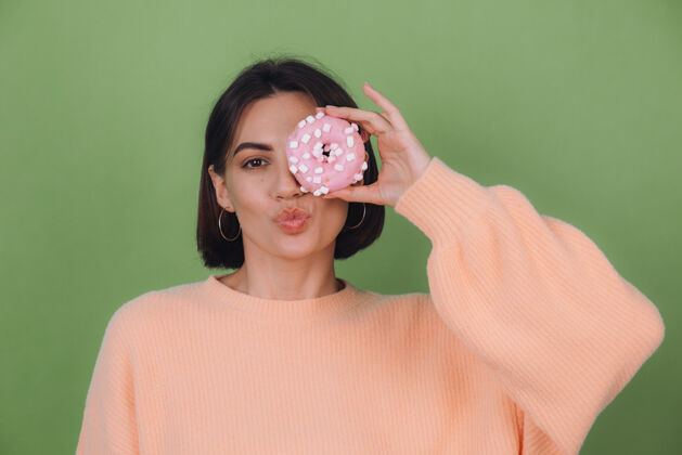 年轻年轻时尚的女士穿着休闲桃色毛衣隔离在绿色橄榄色的墙壁上送上亲吻与粉色的甜甜圈复制空间时尚甜甜圈乐趣