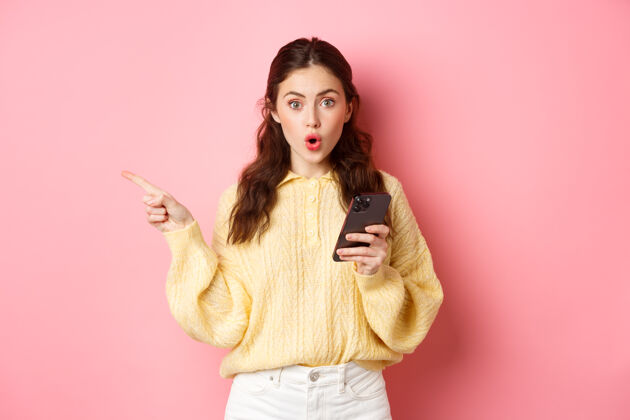 时尚惊讶的年轻漂亮女孩喘着粗气说哇 手指指着左边的版面 拿着手机 站在粉红色的墙上科技手机手机