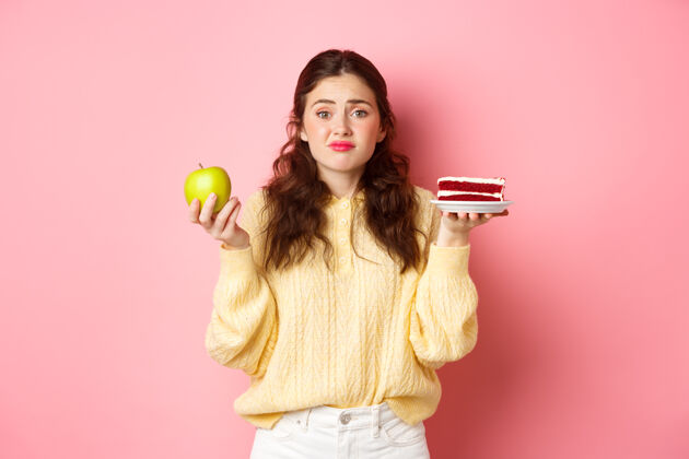 手势年轻可爱的女人在美味的蛋糕甜点和绿色健康的苹果之间犹豫不决 正在节食 犹豫不决地站在粉红色的墙上年轻女人休闲