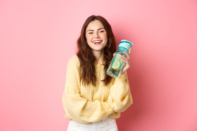积极保持水分面带微笑的健康女孩拿着柠檬水瓶 面带微笑地看着镜头 站在粉色工作室的墙上女孩女人乐观
