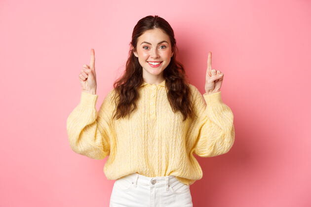 休闲一个漂亮的白人年轻女子手指向上 展示广告 对着镜头微笑 站在粉红色的墙上候选人快乐指向