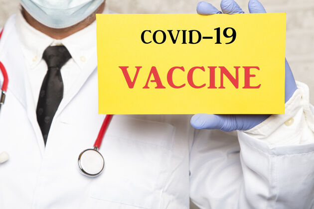 面罩一个戴着手套的男医生拿着一个带有文字covid-19疫苗标志的特写镜头支持室内纸板