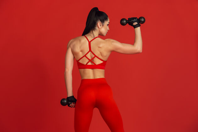 颜色美丽的年轻女运动员在红工作室的墙上练习举重室内运动服