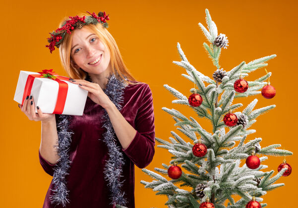花圈站在圣诞树旁的年轻漂亮女孩 身穿红色连衣裙 脖子上戴着花环 手里拿着一个孤立在橙色墙上的礼盒请靠近戴着站着