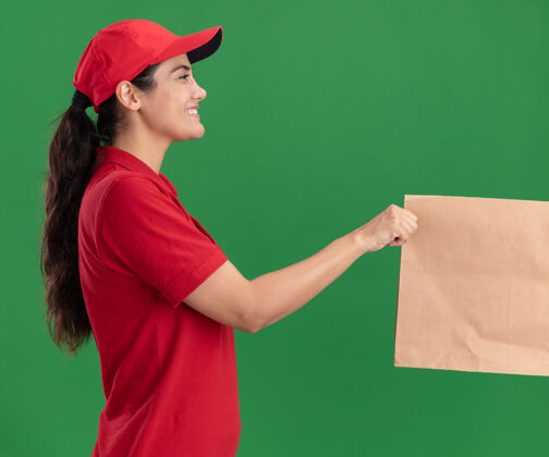 衣服微笑着站在侧视图年轻的送货女孩穿着制服和帽子给纸食品包装客户隔离在绿色的墙壁上给予人客户