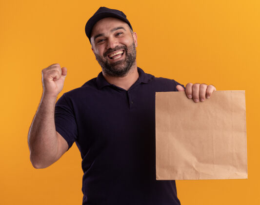 微笑微笑着的中年送货员身着制服 戴着帽子 手里拿着纸食品包 在黄色的墙上显示“是”的手势表情制服帽子